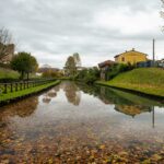 Peaceful autumn canal scene with fallen leaves in Cittadella, Veneto, Italy reflecting cloudy skies.
